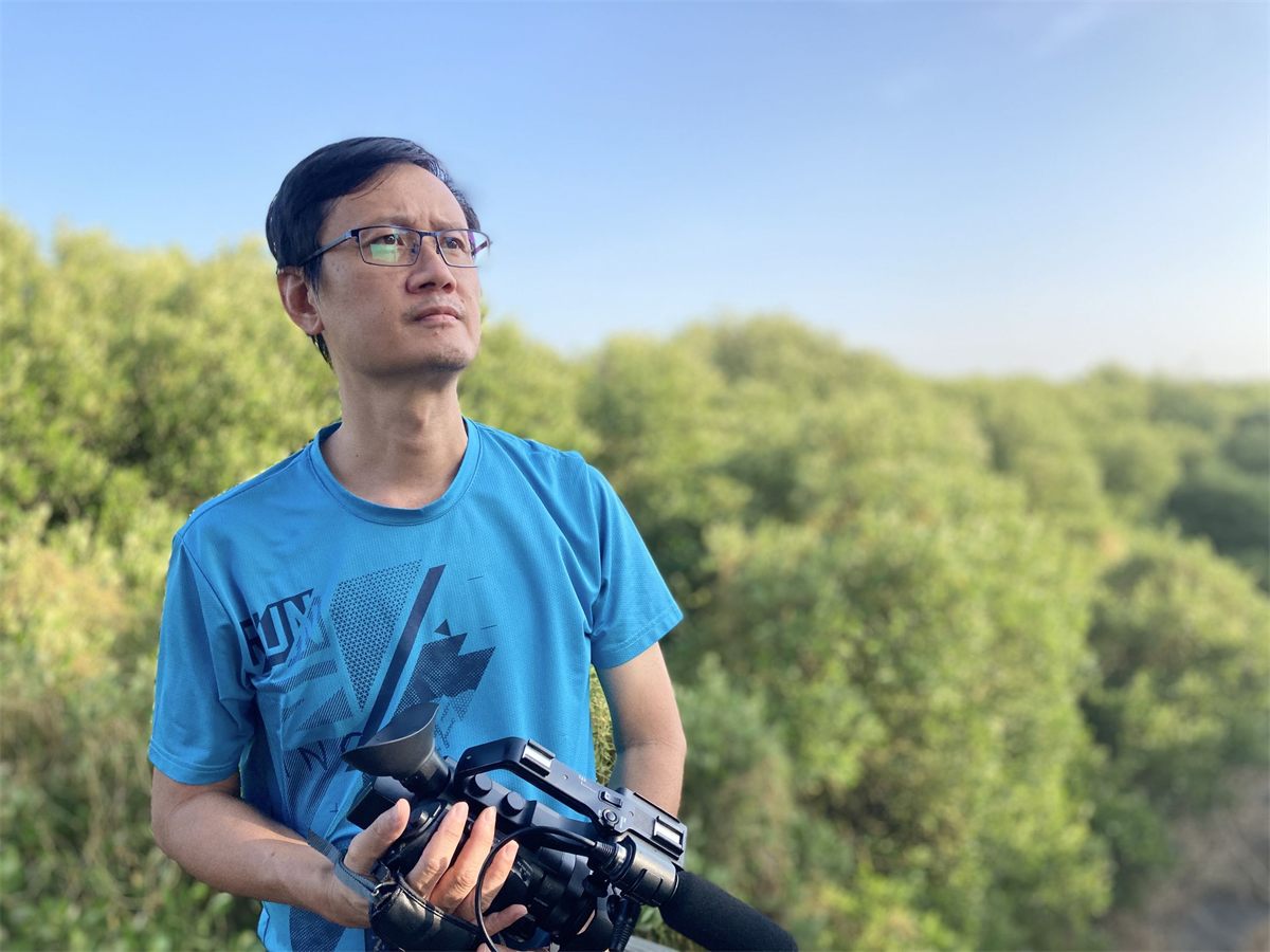 導演吳平海手持攝影機站在綠色樹叢前，望向遠方